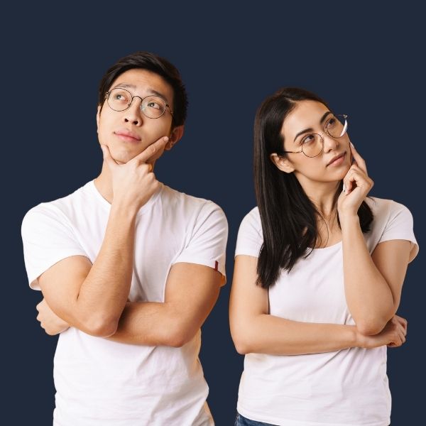 A young couple both wearing glasses and white t-shirts stand against a dark navy background, each with a hand raised to their chin in a thoughtful, contemplative pose.