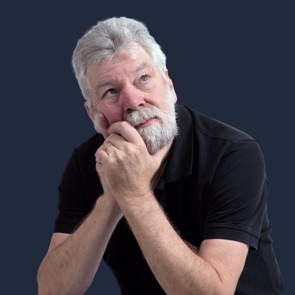 An older man with white hair and a grey beard sits against a dark navy background, hands clasped near his chin, gazing upward with a reflective expression.