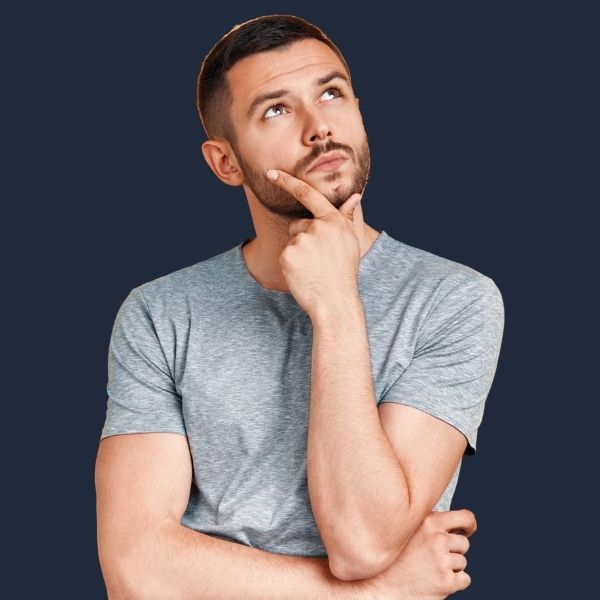 A man in a light denim shirt stands against a dark navy background, one hand resting on the back of his head while he looks upward with a thoughtful, curious expression.